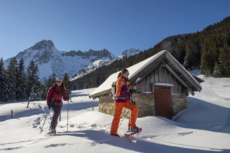 Deux personnes progressent en raquettes dans la neige devant un chalet et les montagnes des Contamines-Montjoie.