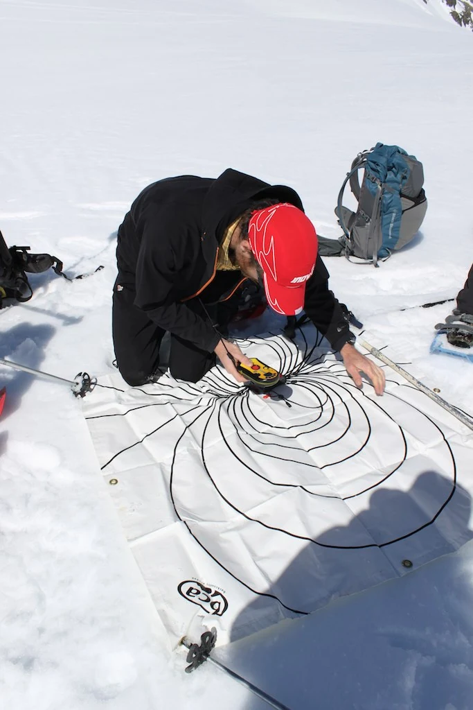 Personne utilisant un DVA sur une grille de recherche dans la neige, avec du matériel de sécurité avalanche.